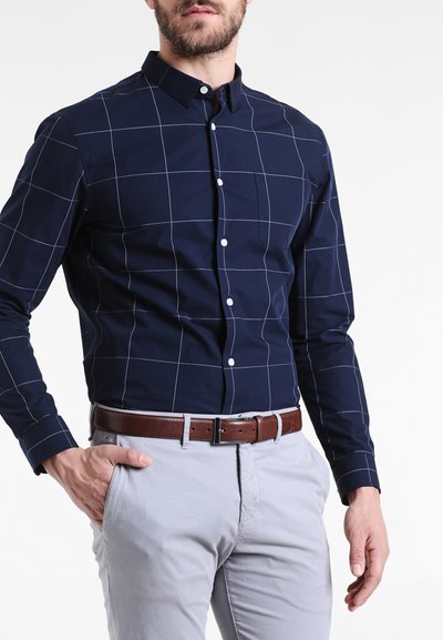 Cămașă albastră închis cu mânecă lungă, cu model de plasă albă, guler cu nasturi și nasturi albi, asortată cu pantaloni gri deschis și o curea maro.