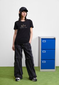 T-shirt noir avec logo doré, pantalons cargo baggy noirs et casquette noire. L'arrière-plan comprend une armoire de classement grise avec des tiroirs bleus sur un sol vert.