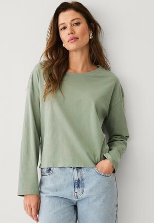 Hellgrünes Langarm-T-Shirt mit rundem Halsausschnitt und überschnittenen Schultern, kombiniert mit hochgeschnittenen blauen Jeans im verwaschenen Muster.