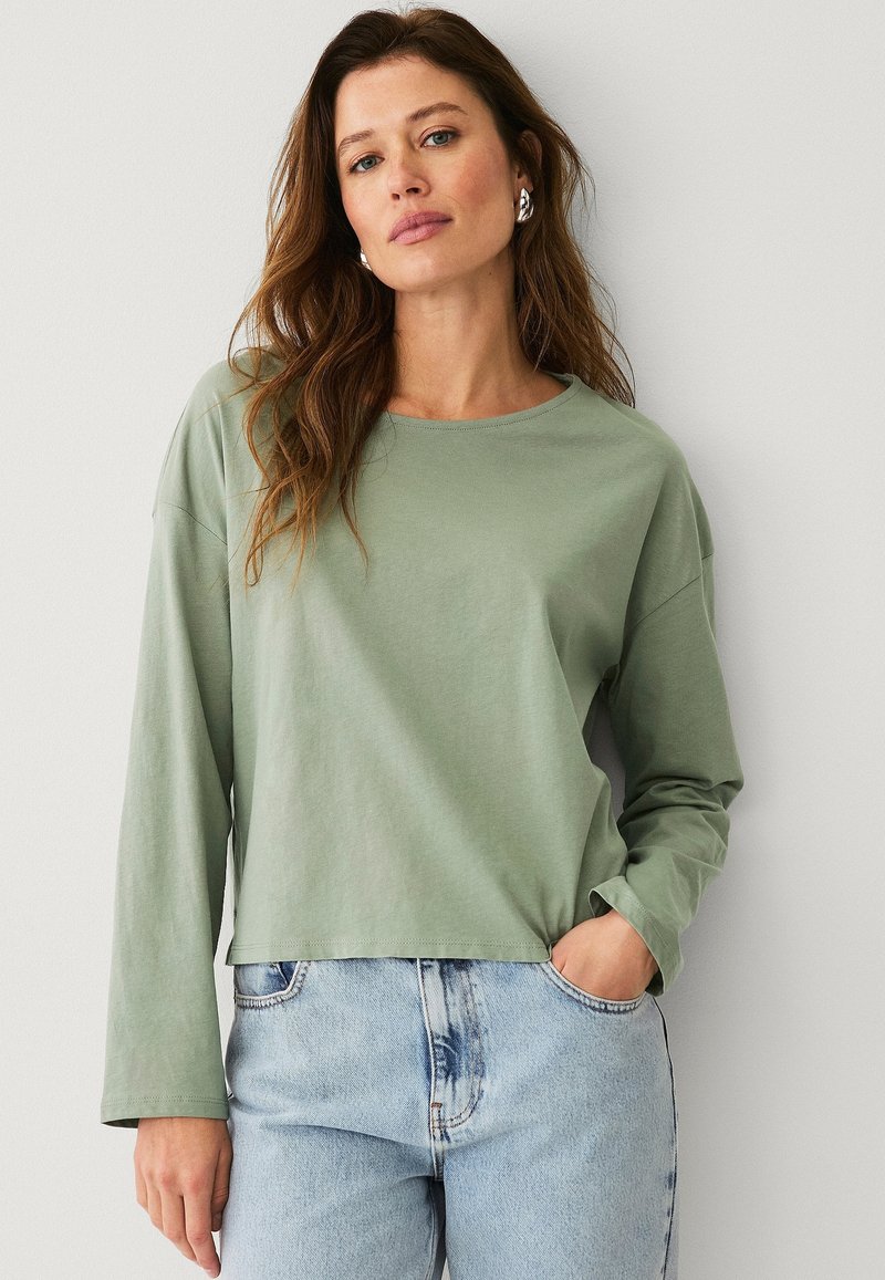 Hellgrünes Langarm-T-Shirt mit rundem Halsausschnitt und überschnittenen Schultern, kombiniert mit hochgeschnittenen blauen Jeans im verwaschenen Muster.