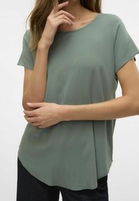 Top de manga corta en verde apagado, hecho de una tela suave. Presenta un cuello redondo y un dobladillo ligeramente curvado, combinado con jeans oscuros.