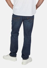 Dunkelblaue Jeans aus Denim mit geradem Schnitt, Gesäßtaschen mit Stichakzenten, getragen über weißen Sneakers. Einfaches weißes T-Shirt ist sichtbar.