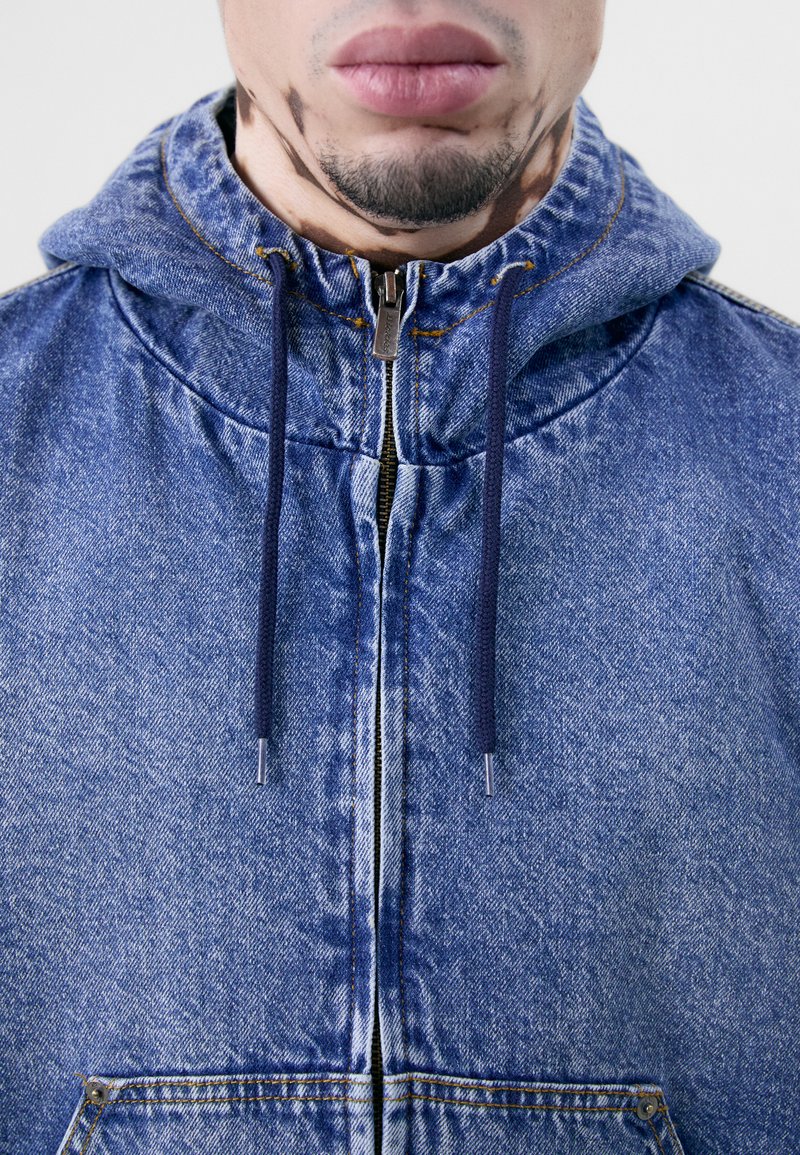 Giacca di jeans azzurra chiara con cappuccio, zip frontale e lacci blu scuro. Presenta una consistenza morbida e dettagli di cucitura visibili.