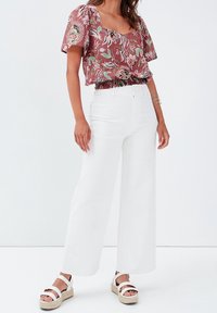 Rotes Crop-Top mit Blumenmuster und kurzen Ärmeln, kombiniert mit weißen hoch taillierten, weit geschnittenen Hosen und weißen Plateausandalen.