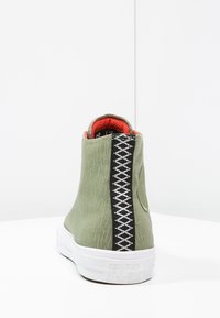 Sneaker alta in tessuto verde con una striscia decorativa a motivi in bianco e nero, suola in gomma bianca e rivestimento interno rosso.