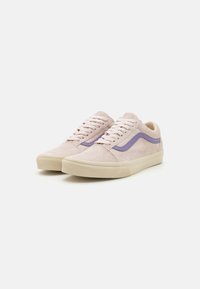 Une paire de baskets en suède rose clair avec des semelles en caoutchouc beige et une bande latérale violette, présentée de biais sur un fond blanc.