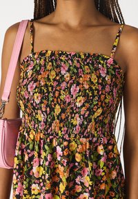 Frau trägt ein schwarzes Blumenkleid mit pinken, orangen und gelben Blumen, dünnen Schulterträgern und einer pinken Handtasche auf ihrer linken Schulter.