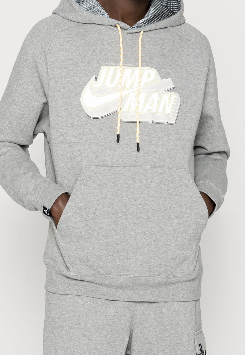 Personne portant un sweat à capuche gris avec l'inscription "Jump Man" et un logo swoosh, les mains dans la poche avant, un pantalon gris assorti visible.