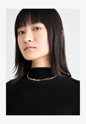 Zlatý retiazkový náhrdelník s oválnymi článkami nosený cez čierny, ryhovaný sveter s vysokým golierom. Textúra je hladká s jemným leskom.