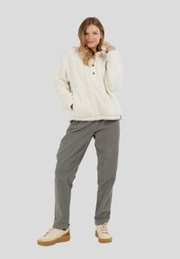 Ung kvinde iført en cremet fleecetrøje, grå bukser og cremede sneakers med gummisåler, stående med den ene hånd i lommen mod en ensfarvet baggrund.