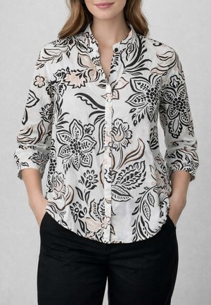Donna che indossa una camicia bianca con bottoni con motivo floreale nero e beige e pantaloni neri, mani nelle tasche, in piedi davanti a uno sfondo grigio.
