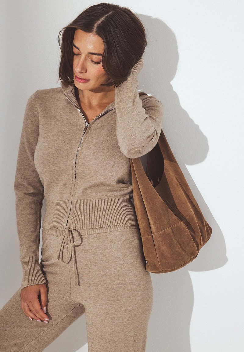 Beige zip-up tröja med ribbad nederkant och matchande byxor, som har dragsko. Åtföljd av en mjuk brun mocka tote-väska.