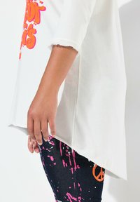 Witte katoenen top met versleten mouwzoom, gecombineerd met donkere stoffen leggings met roze en oranje abstracte patronen.