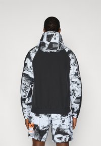 Κουκούλα μπουφάν με μαύρο σώμα και μοτίβα σε ασπρόμαυρα μανίκια, χαλαρή εφαρμογή και ελαστική ζώνη στη μέση. Τα σχέδια είναι αφηρημένα και γωνιώδη.