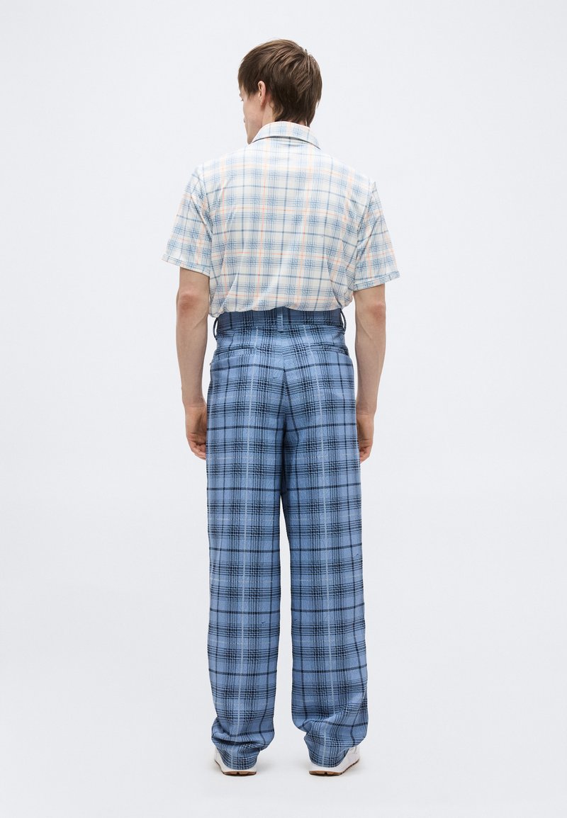 Hombre de espaldas frente a un fondo blanco, vistiendo una camisa de manga corta a cuadros clara y pantalones anchos de pierna ancha de cuadros azules de cintura alta, con zapatos blancos.