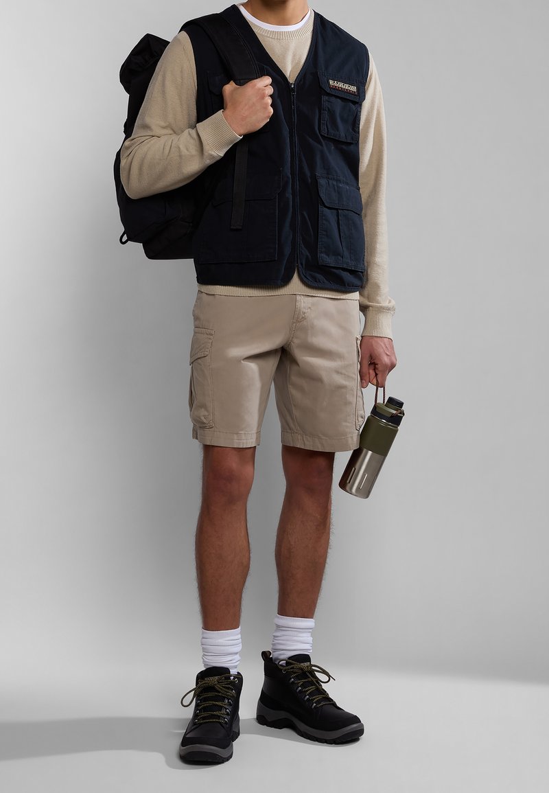 Chaleco navy con múltiples bolsillos sobre un suéter beige, combinado con pantalones cortos de cargo beige. Botines negros y una botella de agua verde completan el conjunto.