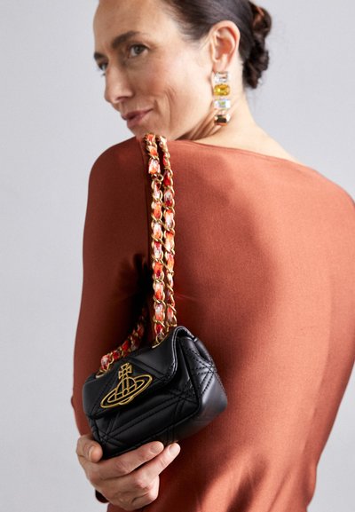 Black quilted handbag with a gold logo, featuring a multicolored braided strap. Woman holds it against a rust-colored top.