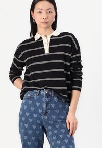 Črno-in-bež progasta polo majica z dolgimi rokavi in rebrastim ovratnikom, v kombinaciji s svetlo modrimi jeans hlačami z vzorcem srčkov.