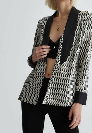 Frau trägt einen schwarz-weiß karierten Blazer mit schwarzen Revers, ein schwarzes Spitzen-Bralette, schwarze Hosen und mehrere goldene Halsketten.