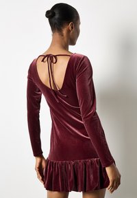 Burgundowa sukienka z weluru z długimi rękawami, niskim dekoltem z tyłu i falbaną u dołu. Gładka faktura i wiązania z tyłu dodają szczegółów.