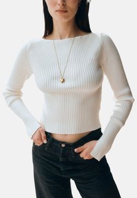 Mujer que lleva un top corto blanco de manga larga acanalado, jeans negros y un collar con un colgante dorado en forma de corazón, de pie con las manos en los bolsillos.