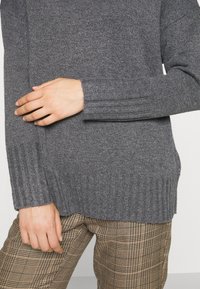 Hände auf der Mittelpartie ruhend, getragen mit einem grauen Rippstrickpullover und beigen Karohosen vor einem neutralen Hintergrund.