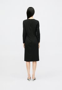 Crna haljina dugih rukava s prilagođenim siluetom, dužine do koljena, glatke teksture i diskretnim detaljem zatvarača na leđima.