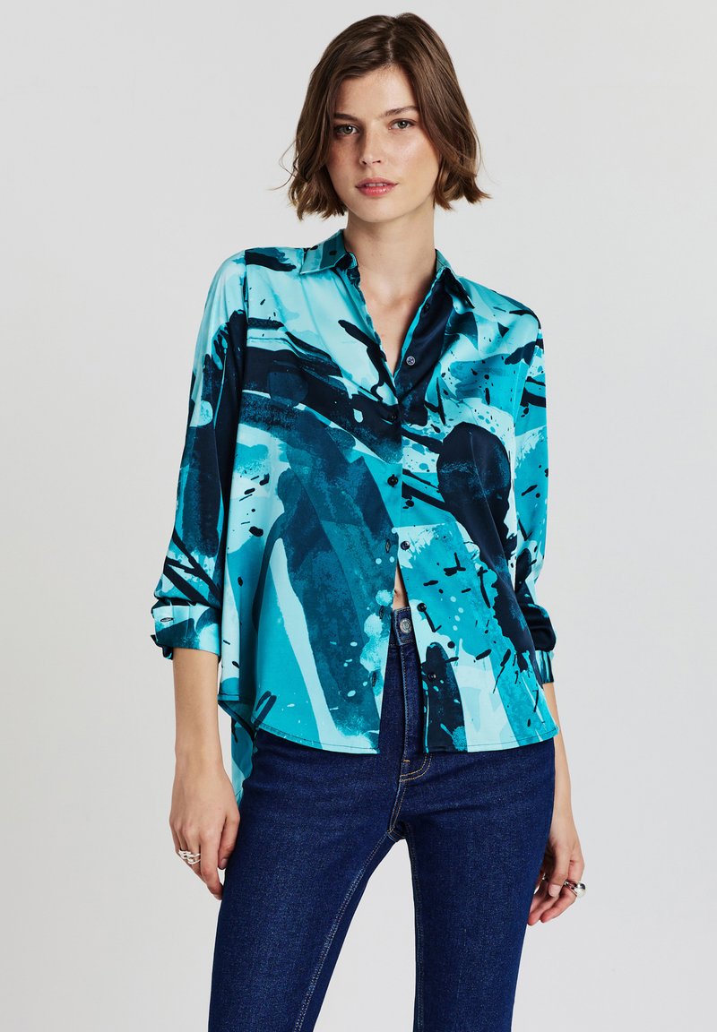 Woman wearing blue abstract patterned long-sleeve button-up shirt and dark blue jeans, standing against plain light background.