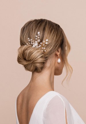 Haare zu einem niedrigen Dutt frisiert, mit einem dekorativen Haarschmuck, der Perlen und floralem Akzenten enthält, vor einem neutralen Hintergrund.