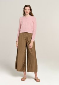 Hellrosa, verkürzter Cardigan mit Rundhalsausschnitt und Knopfverschluss, kombiniert mit olivbraunen, weit geschnittenen Hosen aus zerknittertem Stoff.