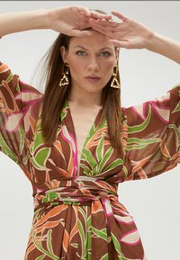 Robe portefeuille marron avec des motifs de feuilles colorées en vert, rose et orange. Elle présente des manches larges et un décolleté plongeant, accentués par une ceinture.