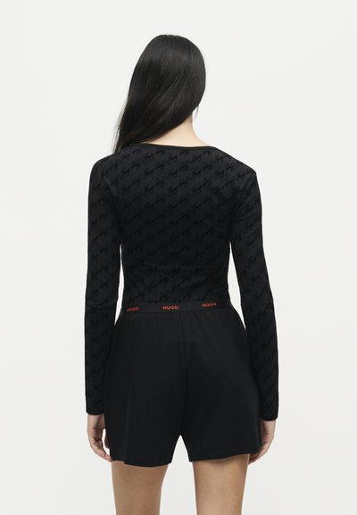 Femme aux longs cheveux foncés portant un haut noir moulant à manches longues et un short noir taille haute avec "HUGO" à la ceinture, vue de dos.