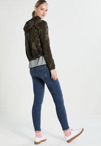 Camo-print cropped jacket in olive green, paired with a gray t-shirt, slim-fit blue jeans, and white sneakers with pink ankle socks.