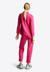 Růžový oblek obsahující přiléhavou blazer a zúžené kalhoty, doplněné šedozelenými teniskami. Tkanina vypadá hladce s pevným designem.