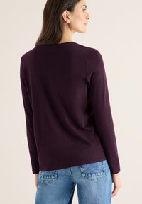 Langærmet top i mørk lilla, lavet af blødt materiale. Har en simpel rund halsudskæring og bæres med blå jeans.