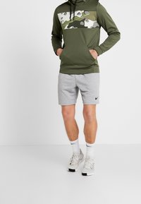 Sudadera con capucha de camuflaje verde con cordón, combinada con pantalones cortos deportivos grises que presentan un logo negro de Nike y zapatillas claras.