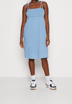 Femme portant une robe sans manches bleu clair avec des liens aux épaules, des chaussettes blanches et des baskets blanches avec des détails bleu foncé, debout sur un fond blanc.
