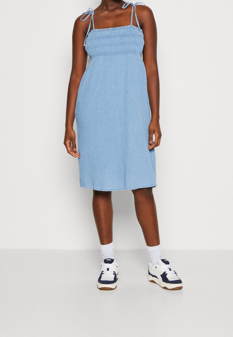 Femme portant une robe sans manches bleu clair avec des liens aux épaules, des chaussettes blanches et des baskets blanches avec des détails bleu foncé, debout sur un fond blanc.