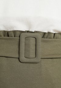 Ceinture en tissu vert olive avec une boucle carrée, présentant une finition texturée et des détails froncés sur le bord supérieur. Chemise blanche au-dessus.
