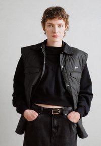 Gilet trapuntato nero sopra un felpa nera, abbinato a jeans neri a vita alta. Presenta una fibbia d'oro e un semplice logo Nike sul gilet.