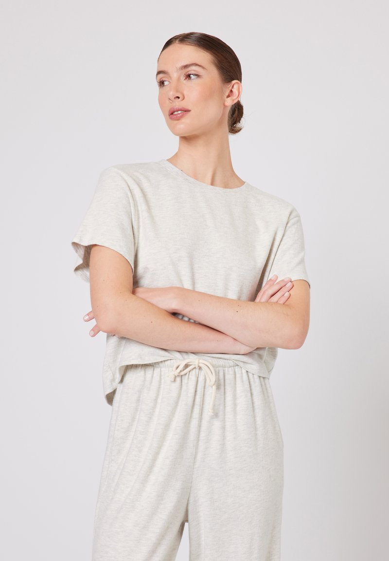 Naine heleroosa lühikeste varrukatega ülaosas ja nööriga pükstes, käed rüpes, vaatab kõrvale ühevärvilise hele tausta ees.