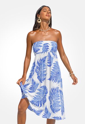 Femme portant une robe blanche sans bretelles avec un imprimé fougère bleu, tenant l'ourlet, les yeux fermés, accessoirisée avec de grandes boucles d'oreilles et un collier de coquillages.