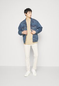 Chaqueta de denim con un lavado azul claro, cierre de botones en el frente, dos bolsillos en el pecho, sobre una camisa beige, combinada con pantalones blancos ajustados y zapatillas.