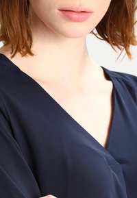 Haut en satin bleu marine avec un design à décolleté en V profond. La texture semble lisse et légèrement réfléchissante, mettant en avant l'élégance.