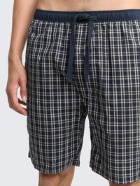 Mand iført marineblå og hvid ternede shorts med elastisk talje og marineblå snor, stående mod en ensfarvet baggrund.