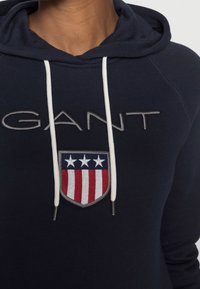 Sweat à capuche bleu marine avec un grand logo blanc "GANT", des cordons blancs, et un écusson en forme de bouclier avec des rayures rouges, blanches et bleues ainsi que des étoiles.