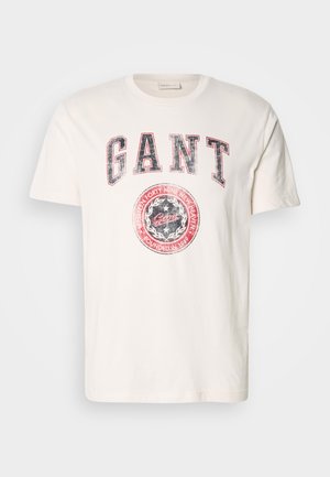 Camiseta de algodón crema con mangas cortas. Presenta un destacado texto en rojo y azul "GANT" y un gráfico circular con "fundada en 1949."