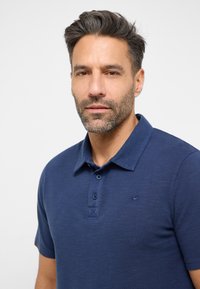 Tamsiai mėlynas polo marškinėliai su tekstūruota medžiaga, trijų mygtukų apykaklė ir mažas logotipas krūtinėje. Trumpi rankovės ir apykaklės dizainas.