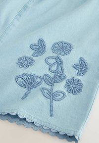 Ljusblå denimtyg med blå broderade blommönster och detaljer med scallopad fåll. Designen inkluderar olika blomformer och blad.