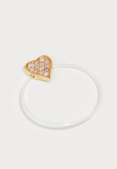 Anillo en forma de corazón dorado con rhinestones rosas en una hendidura en el centro, montado sobre una banda transparente. Textura suave, diseño minimalista.
